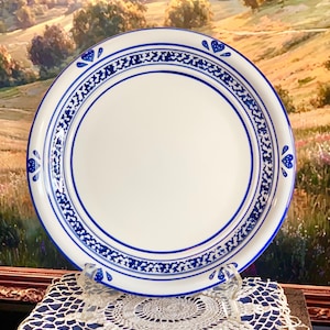Può includere: Un piatto rotondo in ceramica bianca con un bordo decorativo blu. Il bordo è ornato da un motivo ripetitivo di piccoli cuori blu e complessi dettagli floreali, incorniciati da linee circolari blu attorno alla parte centrale.