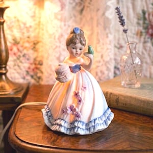 May include: A porcelain figurine lamp shaped like a person with brown hair in an updo, wearing a white dress with blue trim and purple floral accents. The figure holds a small bird on one hand and a small cage in the other, emitting a warm glow.