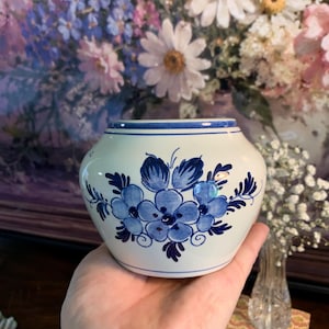May include: A small, round, cream-colored ceramic bowl featuring a hand-painted blue floral pattern on the front. The bowl has a smooth, glossy finish with a blue rim and a thin blue horizontal line encircling the upper portion of the vessel.
