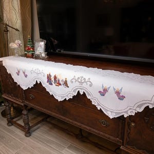 May include: A white rectangular table runner featuring intricate cutwork lace edges and embroidered motifs. The design includes a central nativity scene with figures in colorful robes and several flying angels with purple and blue wings.