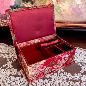 May include: A rectangular jewelry box featuring a red fabric exterior with a floral pattern in shades of cream, green, and gold. The interior is lined with solid red fabric and includes a ring roll and two open compartments for storage.