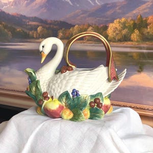 Puede incluir: Una tetera de cerámica con forma de cisne que presenta un cuerpo blanco, un asa curva dorada y roja, y una base decorada con hojas verdes esculpidas, manzanas rojas y pequeñas bayas azules. El cisne tiene un cuello largo y un pico amarillo.