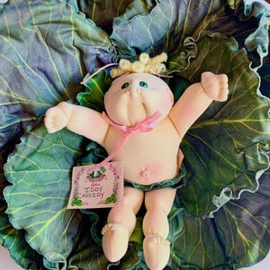 May include: A Cabbage Patch Kids doll with light skin, green eyes, and blonde yarn hair styled in curls. The doll wears a pink ribbon around its neck and has a tag attached that reads IDDY CASSIDY. It is positioned on a large, green cabbage leaf.