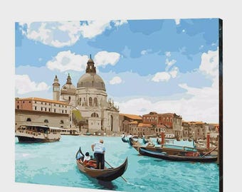 May include: A painting of a Venetian canal scene featuring a gondolier in a white shirt and dark trousers rowing a black gondola. In the background, a large domed stone church stands near terracotta-colored buildings under a bright blue sky.