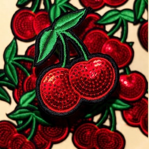 May include: A collection of cherry-shaped embroidered patches featuring bright red sequined fruit and vibrant green stitched leaves. Each patch is outlined with a thick black border, creating a distinct contrast against the shiny red sequins.