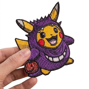 Puede incluir: Un parche bordado que muestra un personaje amarillo con mejillas rojas vestido con un disfraz morado de orejas puntiagudas y una amplia sonrisa dentada. El personaje sostiene un pequeño cubo de calabaza naranja, con detalles en negro y amarillo.