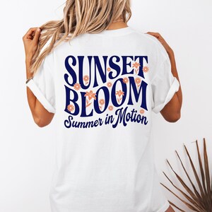 May include: A white short-sleeved t-shirt featuring a large graphic on the back with the dark blue text SUNSET BLOOM and the script text Summer in Motion, accented by small orange flower illustrations integrated into the lettering.