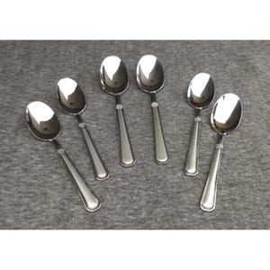 May include: A set of six polished stainless steel spoons arranged in two rows. Each spoon features a smooth, reflective oval bowl and a straight, flat handle with a subtle decorative band of three horizontal lines near the neck of the spoon.