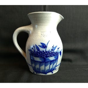 May include: A light gray stoneware pitcher featuring a prominent blue embossed design of a basket filled with berries and leaves. The vessel has a wide, flared rim, a sturdy side handle, and a small stamped mark near the top of the body.