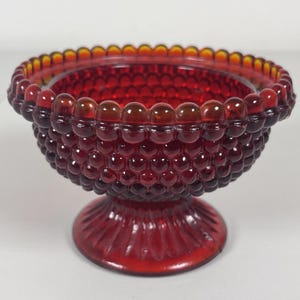 Puede incluir: Un cuenco de cristal rojo intenso con una textura de puntos en relieve por toda la superficie exterior. El cuenco tiene un borde festoneado y se apoya sobre una base circular estriada. El cristal es translúcido y deja pasar la luz.