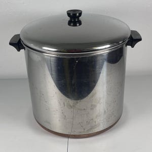 Puede incluir: Una olla grande y cilíndrica de acero inoxidable con acabado pulido y base de color cobre. Cuenta con dos asas laterales negras y un pomo negro a juego en el centro de la tapa circular. La superficie metálica muestra ligeras marcas de uso.
