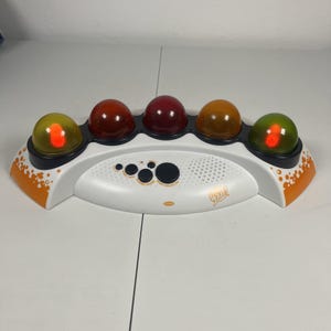 Op de afbeelding: Een witte elektronische spelconsole met vijf doorschijnende, koepelvormige knoppen in geel, rood, donkerrood, oranje en groen. De basis bevat zwarte bedieningsknoppen, een luidsprekerrooster en de tekst "What?" in het oranje afgedrukt.