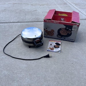 May include: A round, chrome-finished Mickey's Waffler appliance with a black handle and power cord, accompanied by its original red and white cardboard packaging box and a printed recipe and instruction booklet featuring waffle illustrations.