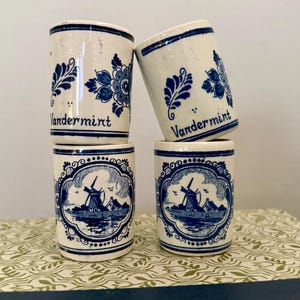 May include: Four cylindrical ceramic shot glasses featuring a white base with blue decorative patterns. Two glasses display the text Vandermint with floral motifs, while the other two feature a detailed blue illustration of a Dutch windmill.