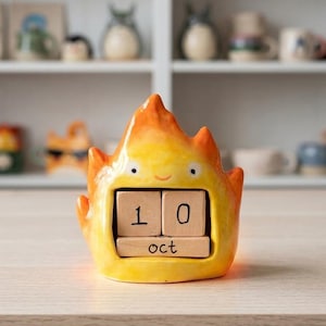 Könnte beinhalten: Ein flammenförmiger ewiger Kalender aus Keramik mit gelb-orangem Farbverlauf. Die Figur hat ein einfaches Gesicht mit zwei Augen und einem kleinen Lächeln. Sie enthält zwei Holzblöcke mit der Zahl 10 und einen Block mit der Aufschrift oct.