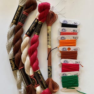 Op de afbeelding: Assortiment van borduurgaren spoelen in verschillende kleuren, waaronder zilver, bruin, rood, oranje en groen. De spoelen zijn geëtiketteerd met DMC-nummers: 310, 351, 740, 400, 321 en 1099.