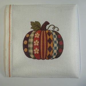 May include: A square piece of white needlepoint canvas featuring a central pumpkin design. The pumpkin is divided into vertical segments with varied patterns including flowers, stripes, diamonds, and checks in shades of orange, red, yellow, and brown.