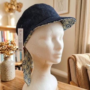 Könnte beinhalten: Eine wendbare Kappe mit einer dunkelblauen Jeans-Außenseite und einem cremefarbenen Innenfutter mit einem sich wiederholenden blauen geometrischen Sternmuster. Die Kappe verfügt über zwei seitliche Bänder und ein weißes Etikett am Schirm.
