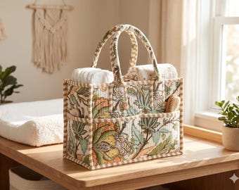 May include: A rectangular quilted fabric storage caddy featuring a floral pattern in shades of green, yellow, and brown with tan striped trim. The caddy has two top handles and multiple exterior pockets containing white diapers and a cloth.