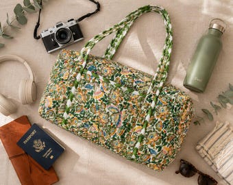 May include: A quilted duffel bag featuring a vibrant floral and butterfly pattern in shades of green, yellow, and orange, with two braided handles. The bag is surrounded by a camera, headphones, a passport, a water bottle, and sunglasses.