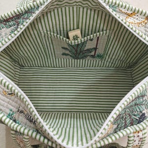 Puede incluir: Un bolso tote acolchado con forro interior de rayas blancas y verdes y un bolsillo interior. El exterior presenta un estampado bot&aacute;nico con hojas verdes y detalles amarillos. Una ficha de mahjong con el car&aacute;cter 發 se encuentra en el interior.