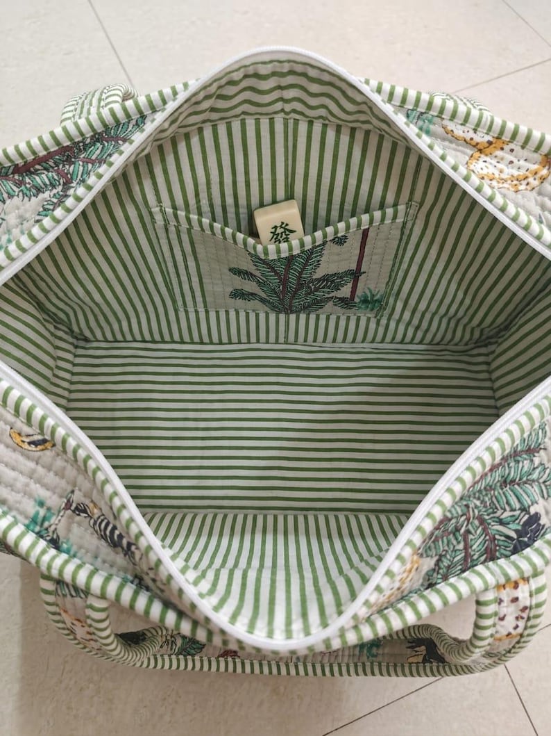 Puede incluir: Un bolso tote acolchado con forro interior de rayas blancas y verdes y un bolsillo interior. El exterior presenta un estampado bot&aacute;nico con hojas verdes y detalles amarillos. Una ficha de mahjong con el car&aacute;cter 發 se encuentra en el interior.