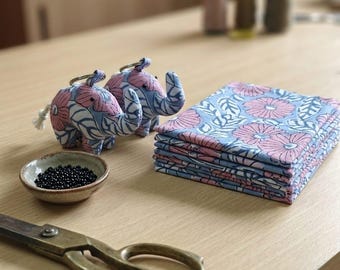 May include: Two stuffed elephant keychains with a blue and pink floral pattern, a stack of folded fabric featuring the same floral print, a small ceramic bowl filled with tiny black beads, and a pair of vintage-style metal sewing scissors.