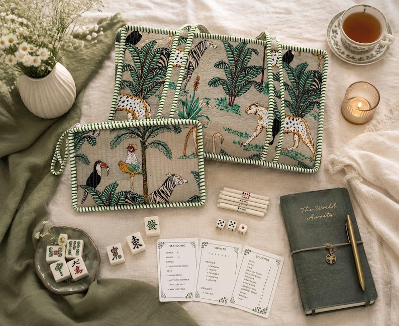 Puede incluir: Un bolso tote rectangular y un estuche con cremallera a juego, ambos de tela beige con un estampado de selva verde con leopardos, cebras y palmeras. El conjunto incluye fichas de mahjong blancas, dados, tarjetas de puntuaci&oacute;n y un diario.