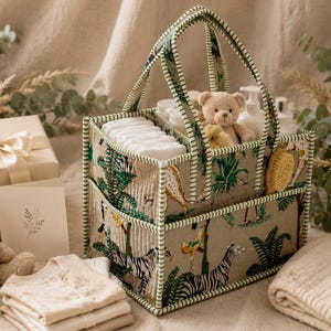 Op de afbeelding: Een rechthoekige stoffen opbergmand met twee handvatten en een groen-wit gestreepte rand. De buitenkant heeft een jungle-print met zebra's, giraffen en tropische planten op een beige achtergrond. De mand bevat witte luiers en een teddybeer.