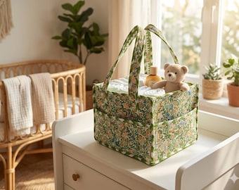 May include: A square fabric storage caddy featuring a green, orange, and white floral pattern with two sturdy handles. The caddy contains white rolled cloths, a small amber-colored bottle, and a light brown plush teddy bear toy.