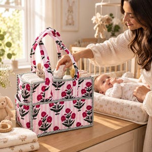Könnte beinhalten: Ein rechteckiger Windel-Organizer aus Stoff mit einem vertikalen Streifenmuster in Rosa und Weiß sowie einem Blumenmuster aus dunkelrosa Blüten und grünen Stielen. Die Tasche hat zwei stabile Griffe und eine schwarz-weiß gestreifte Kante.