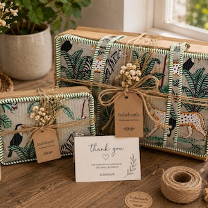 Puede incluir: Dos cajas rectangulares de tela acolchada con estampado de selva con tigres, leopardos y tucanes, acentuadas con ribetes a rayas verdes. Cada caja est&aacute; atada con cordel y una etiqueta de papel marr&oacute;n con la marca Indiehaath.
