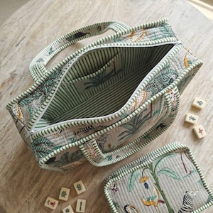 Puede incluir: Un bolso de tela acolchada y un estuche rectangular a juego con cremallera, con un estampado de selva que incluye cebras, tucanes y plantas tropicales sobre un fondo gris claro. El bolso tiene un forro de rayas verdes y blancas.