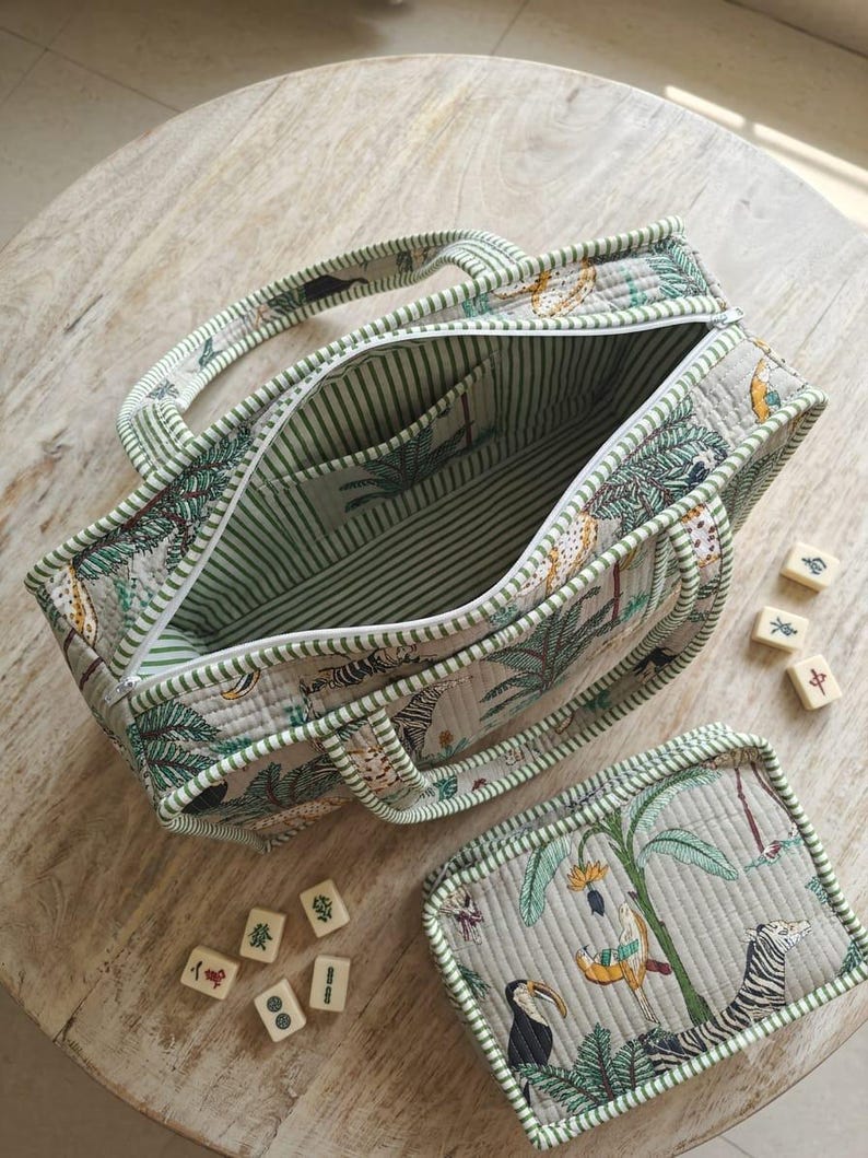 Puede incluir: Un bolso de tela acolchada y un estuche rectangular a juego con cremallera, con un estampado de selva que incluye cebras, tucanes y plantas tropicales sobre un fondo gris claro. El bolso tiene un forro de rayas verdes y blancas.