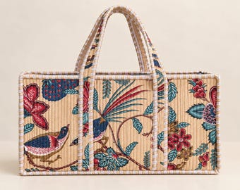 Puede incluir: Un bolso tote acolchado rectangular con fondo beige y un patrón repetitivo de pájaros coloridos, flores rojas y follaje azul. El bolso tiene dos asas a juego y está rematado con un borde de tela a rayas blancas y azules.