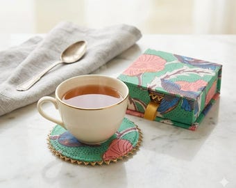 May include: A cream-colored ceramic teacup with a gold rim sits on a round, green fabric coaster featuring a pink and blue floral pattern and a gold scalloped edge. Beside it is a matching square fabric box with a gold ribbon pull tab.