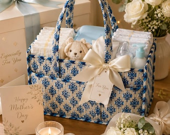 May include: A quilted fabric storage caddy featuring a blue and white floral pattern, filled with baby supplies including diapers, a small plush bear, and a bottle. A cream-colored ribbon bow is tied to the handle with a tag reading For You.