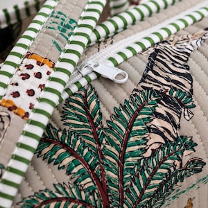 Puede incluir: Un bolso de tela beige acolchada con un patr&oacute;n estampado de tigres y palmeras. El bolso incluye un cierre de cremallera blanco y asas decoradas con un patr&oacute;n de rayas horizontales verdes y blancas, mostrando costuras detalladas en todo el dise&ntilde;o.