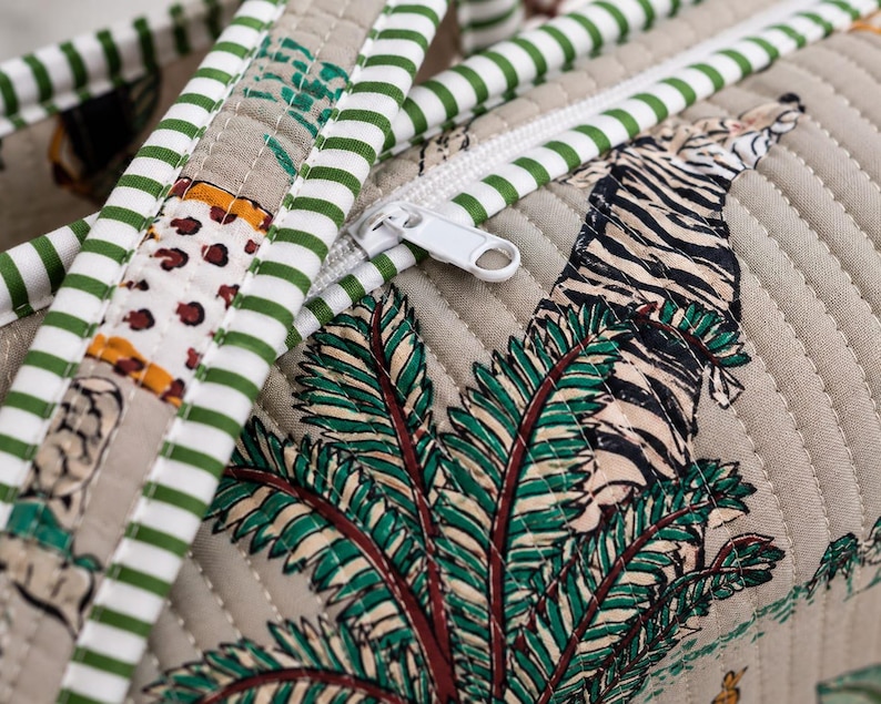Puede incluir: Un bolso de tela beige acolchada con un patr&oacute;n estampado de tigres y palmeras. El bolso incluye un cierre de cremallera blanco y asas decoradas con un patr&oacute;n de rayas horizontales verdes y blancas, mostrando costuras detalladas en todo el dise&ntilde;o.