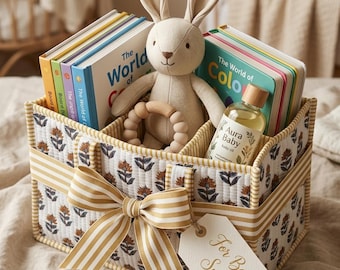 May include: A quilted fabric storage caddy with a floral pattern, featuring a large striped bow and a gift tag labeled For Baby Smith. Inside are several board books, a plush rabbit toy, a wooden teething ring, and a bottle of Aura Baby oil.