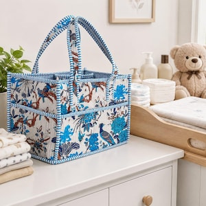 Puede incluir: Una cesta de almacenamiento rectangular de tela con base blanca y un estampado botánico azul y marrón de pájaros y flores. La cesta tiene dos asas verticales y un borde a rayas azules y blancas, con múltiples bolsillos exteriores.