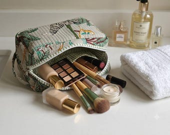 May include: A quilted fabric cosmetic bag featuring a green and white jungle-inspired print with tigers and palm leaves. The open bag contains various makeup items, including a rectangular eyeshadow palette, foundation bottles, and makeup brushes.