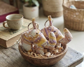 May include: Two stuffed elephant keychains made from fabric with a yellow, purple, and white floral pattern. Each elephant features a small metal keyring attached to the top, a rounded body, and a trunk curled upward, resting inside a wooden bowl.