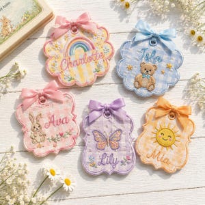 May include: Five scalloped fabric ornaments with gingham patterns and satin bows. The ornaments feature embroidered names and motifs: Charlotte with a rainbow, Isla with a teddy bear, Ava with a rabbit, Lily with a butterfly, and Mia with a sun.