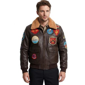 May include: A dark brown leather bomber jacket featuring a tan faux fur collar, a front zip fastening, and ribbed cuffs. The jacket is adorned with multiple colourful embroidered military-style patches on the chest and sleeves, including text.