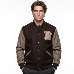 Könnte beinhalten: Eine dunkelbraune Collegejacke mit hellbraunen Ärmeln sowie geripptem Kragen, Bündchen und Saum. Die Jacke verfügt über eine Druckknopfleiste, zwei Seitentaschen mit hellbrauner Einfassung und einen großen, hellbraunen Buchstaben B auf der Brust.