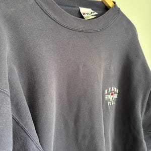 May include: A navy blue crewneck sweatshirt featuring a small embroidered U.S. Open logo on the chest. The logo includes the text U.S. Open, New York, and the Fila brand name with a red and blue graphic element. A white Fila brand tag is visible at the neck.