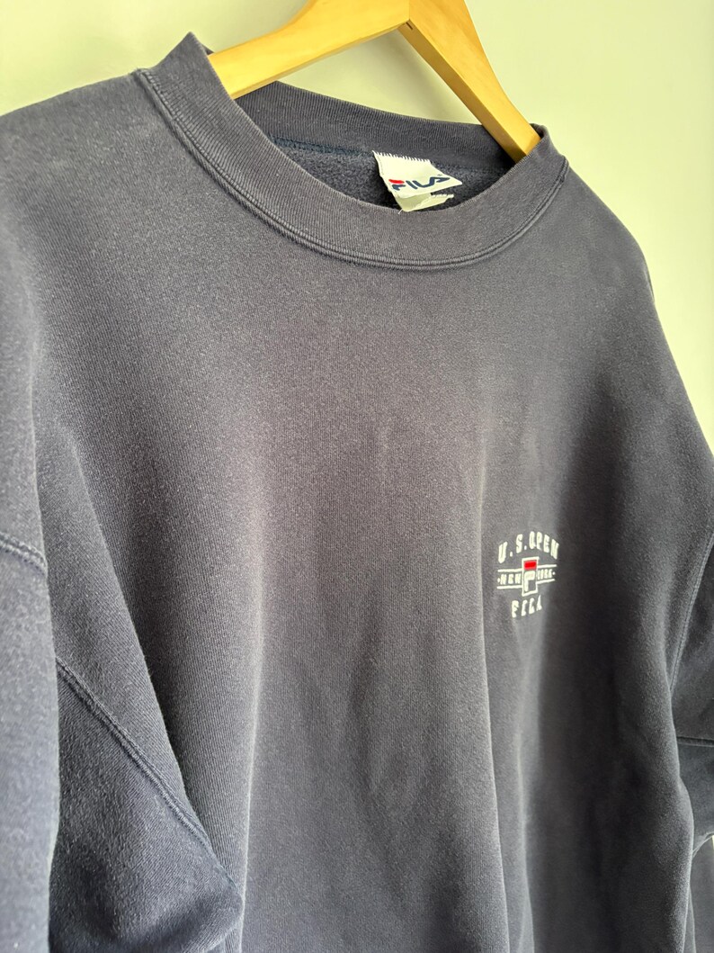 May include: A navy blue crewneck sweatshirt featuring a small embroidered U.S. Open logo on the chest. The logo includes the text U.S. Open, New York, and the Fila brand name with a red and blue graphic element. A white Fila brand tag is visible at the neck.
