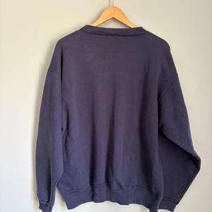 May include: A solid navy blue long-sleeve sweatshirt featuring a crew neckline, ribbed cuffs, and a ribbed hem. The garment is displayed on a wooden hanger against a plain background, showing the back side of the pullover with simple stitching.