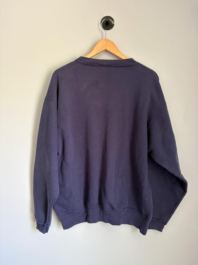 May include: A solid navy blue long-sleeve sweatshirt featuring a crew neckline, ribbed cuffs, and a ribbed hem. The garment is displayed on a wooden hanger against a plain background, showing the back side of the pullover with simple stitching.
